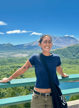 Woman wearing navy blue shirt with green pants and glasses posing in front of mountain view point.