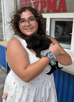 Woman in white dress wearing glasses with curling brown hair, holding a puppy in arms.