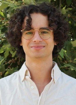 a student with short curly hair wears glasses and a white collared shirt while standing in front of a bush
