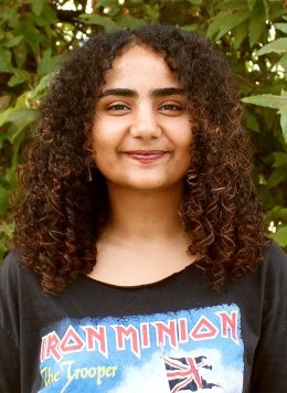 a student with curly long brown hair wears a iron minion tshirt while posing in front of a bush
