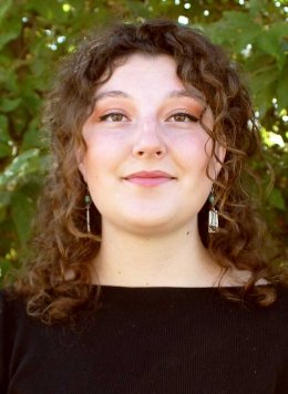 a student with long curly brown hair wears a black shirt while standing in front of a large bush