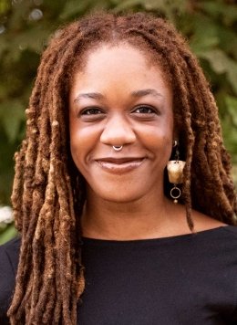portrait of Chi Adi wearing a black top and long earrings, standing in front of an outdoor plant
