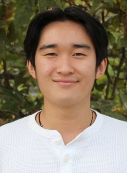 a student with short black hair wears a white henley shirt while standing in front of outdoor bushes