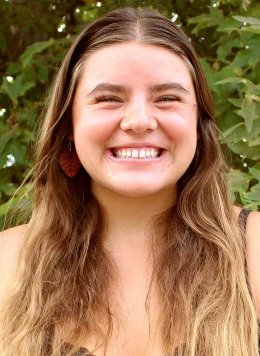 a student with long brown hair stands in front of an outdoor plant