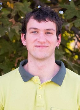 head shot of Raffi Charkoudian Rogers wearing a yellow shirt, standing in front of an outside plant