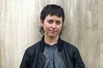 a person with short brown hair wearing a black silk blouse and black bomber jacket stands in front of a concrete wall