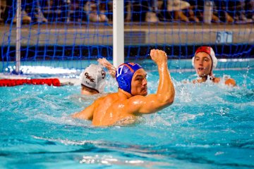 A member of the Pomona-Pitzer men’s water polo team wearing a blue swim cap holds up a fist victoriously in the pool.