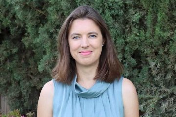 a woman with shoulder-length brown hair stands in front of a bush