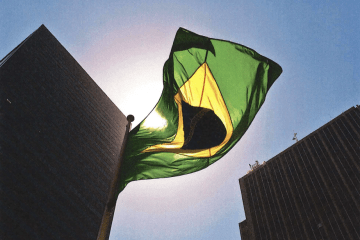 a Brazilian flag with high rises and sky behind it