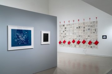 Art gallery with a blue and black photos in white frames and a large newsprint work of art with red folders hanging from the word SECURITY
