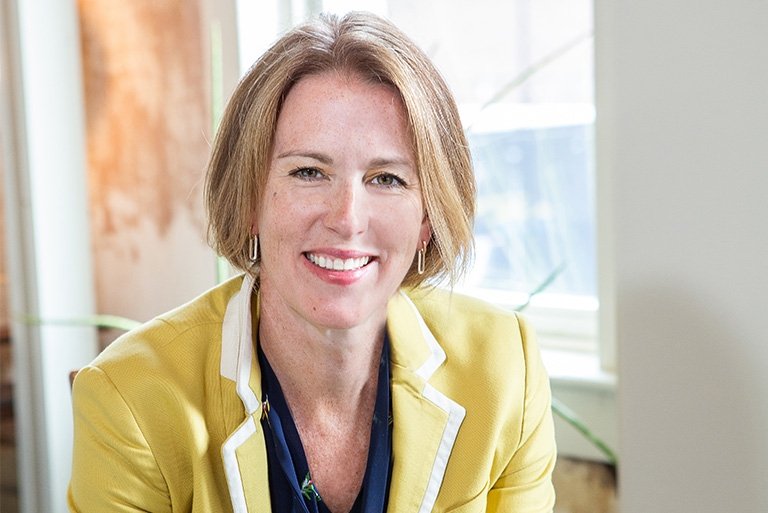 Tricia Milford-Hoyt has straight blond hair in a bob cut and wears a yellow blazer over a blue shirt.