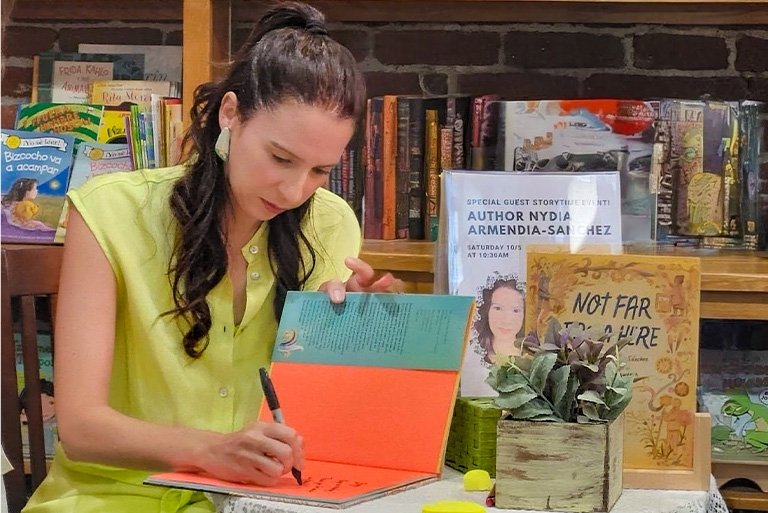 Nydia Armendia-Sanchez has long dark hair and wears a pale yellow top while signing a children’s book.