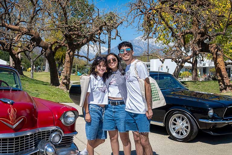 three students pose in front of two classic cars parked near the mounds