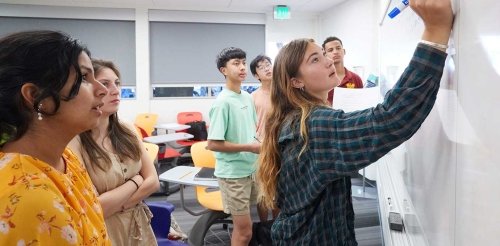 student using expo marker writing on the classroom board while other students watch