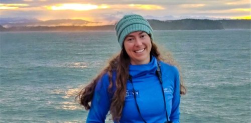 a woman with long brown hair wears a blue stocking hat and puffer jacket while posing in front of ocean water