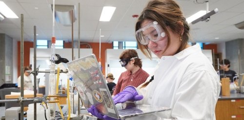 a student wears googles and peers at a laptop while working in a chemistry lab