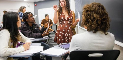 a student in a polka dot dress stands at a blackboard while working with a group of other students sitting at desks