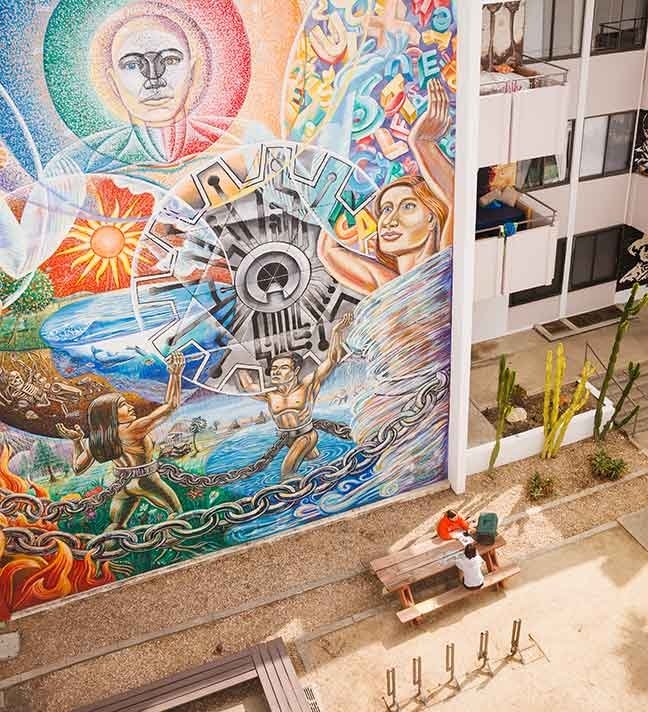 two students sit at a picnic table with a large colorful mural behind them
