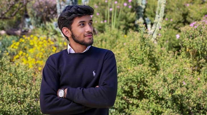 Isa Iqbal wears a dark blue sweater and stands with his arm cross in front of shrubbery