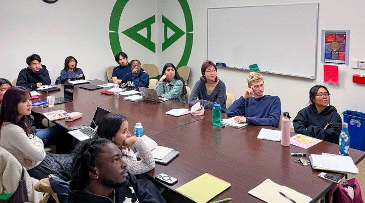 Students in the Trans-Pacific Japan class sit at a long wood table in a classroom while they look at something off-screen.
