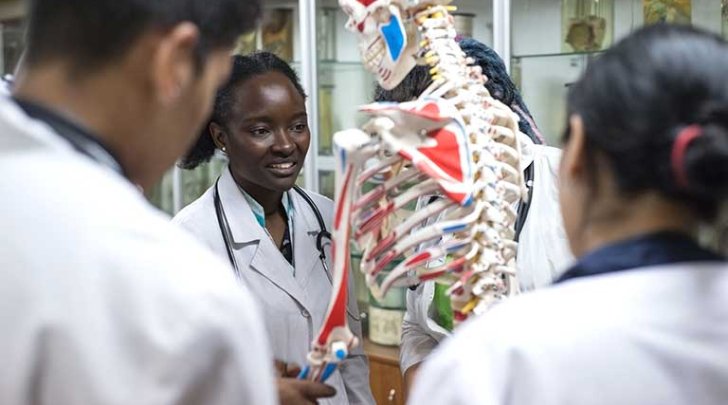 students in white coats surround an educational skeleton in a classroom