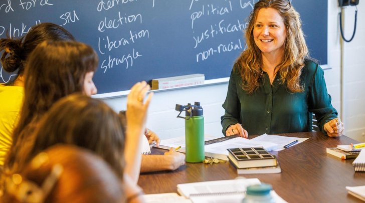 Professor Harmony O'Rourke sits at a table with students with a blackboard in the background