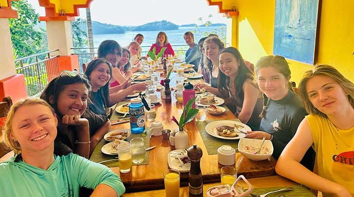 several students sit around a table in a yellow room with a view of the ocean in the background