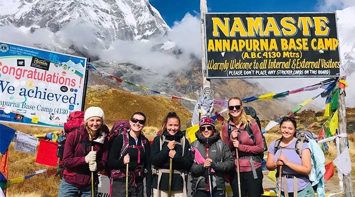 a group of student hikers pose in front of the annapurna base camp sign with snowy mountains in the background