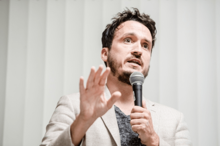 Michael Ceraso speaks on a microphone, wears a white blazer, and has short dark hair and beard.