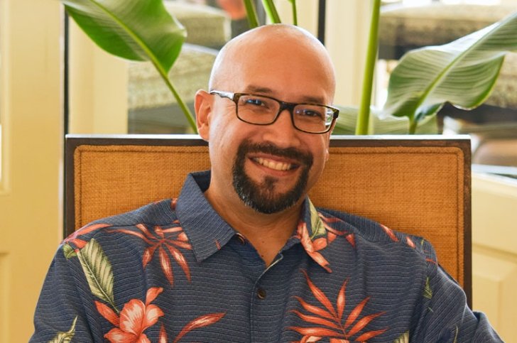 Professor Adrian Pantoja is bald, has a close-cropped beard, and wears black-rimmed glasses and a blue collared shirt with floral print.