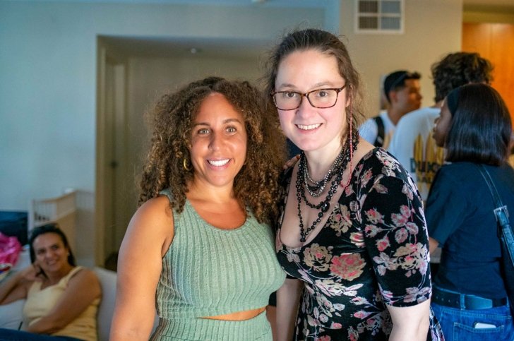 Professor Fely Catan wears teal outfit and Professor Amanda Louise Johnson wears a floral dress inside a faculty apartment.