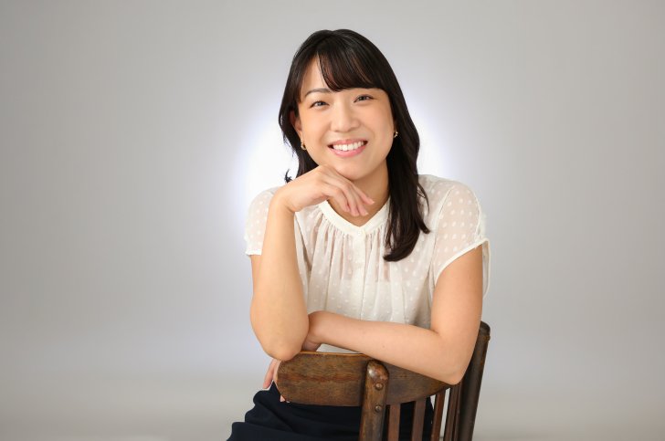 Mao Kobayahsi sits on a wood chair, facing toward the camera