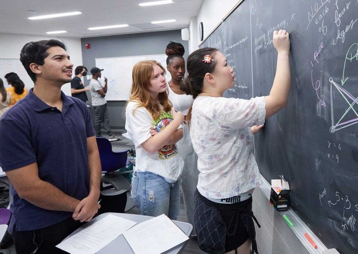 Student writing with chalk on blackboard while others watch