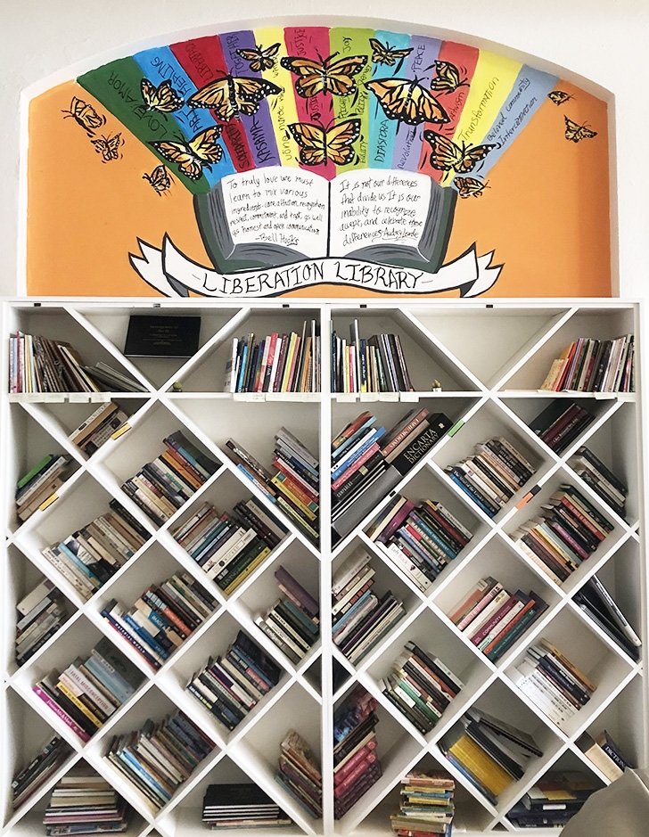 a tall white bookshelf is against the wall. Above it is bright mural with butterflies and rainbows with the words "libration library"