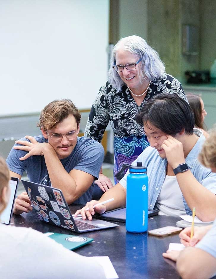 two students sit at a desk while a professor looks over their shoulders at a laptop screen