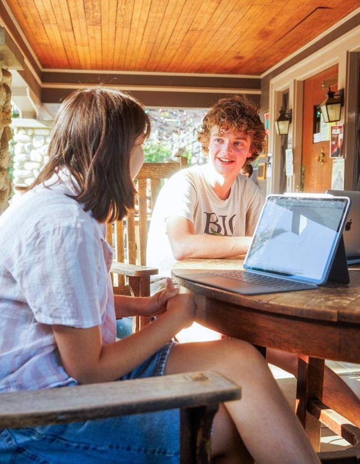 two students sit at a table on the grove house porch in front of computers