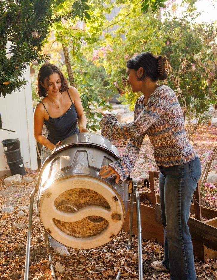 two student spin a drum compost container in the student garden