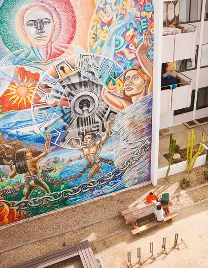 two students sit at a picnic table with a large colorful mural behind them