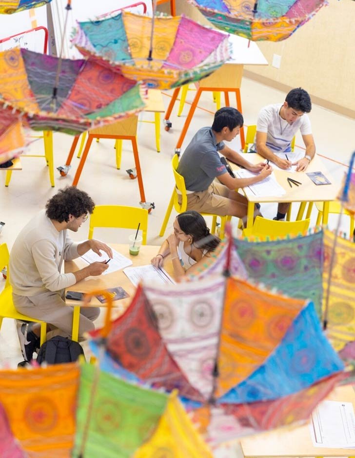 four students sit at tables doing work in a room decorated with bright umbrellas