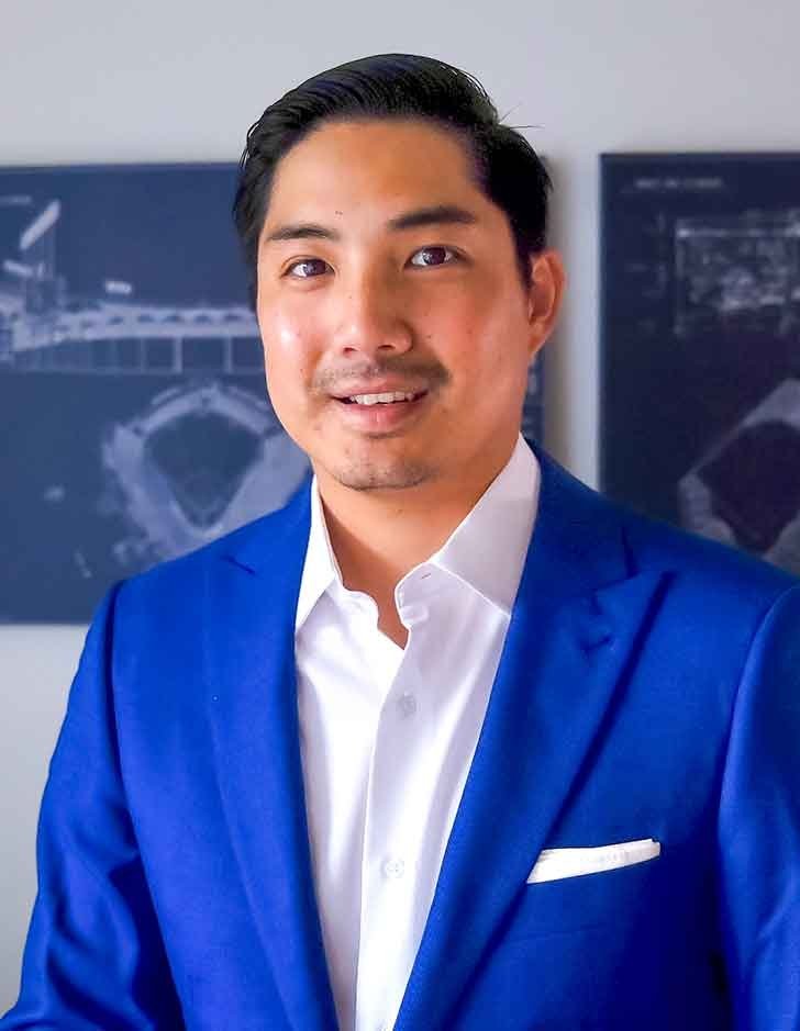 James Kang wears a blue blazer and white shirt while standing in front of stadium blueprints on a white wall