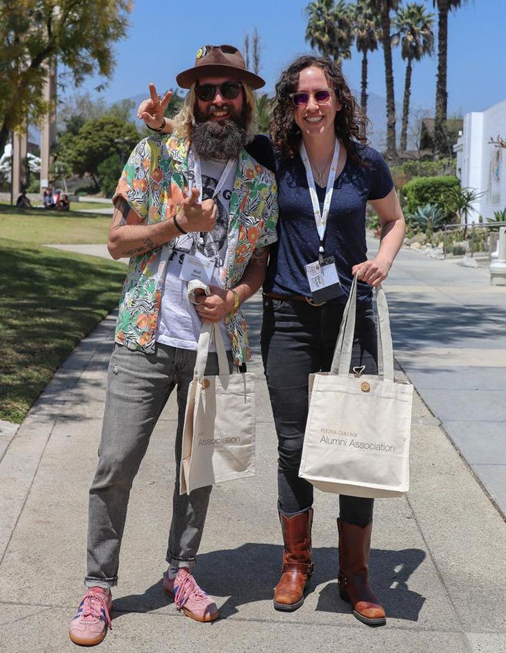 two pitzer alums pose near the mounds while holding alumni association tote bags