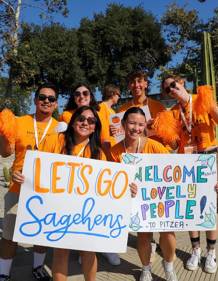 students in orange shirts hold "let's go pitzer" and "welcome lovely people" signs at move in day