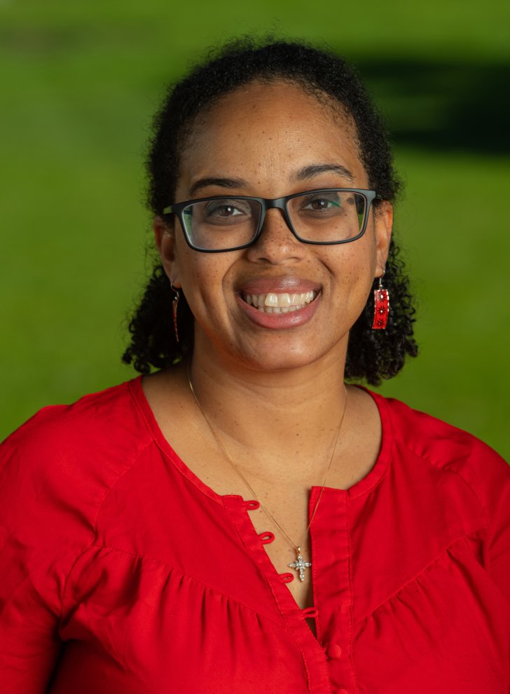 Woman wearing glasses and wearing red earrings and red blouse, standing in front of greenery.