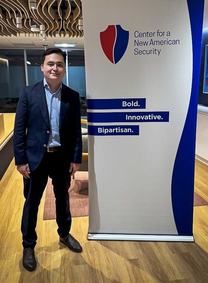 A student with short brown hair , wearing a button shirt and blazer stands next to a sign that says "Center for a New American Security"