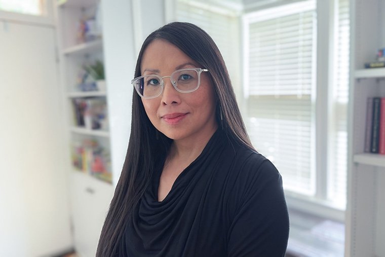 portrait of woman with long brown hair and glasses with a window and bookshelf behind her