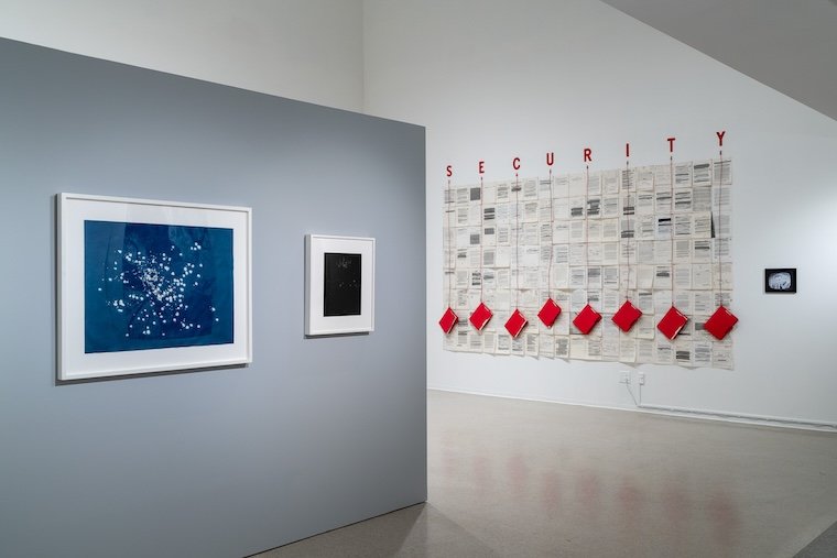 Art gallery with a blue and black photos in white frames and a large newsprint work of art with red folders hanging from the word SECURITY