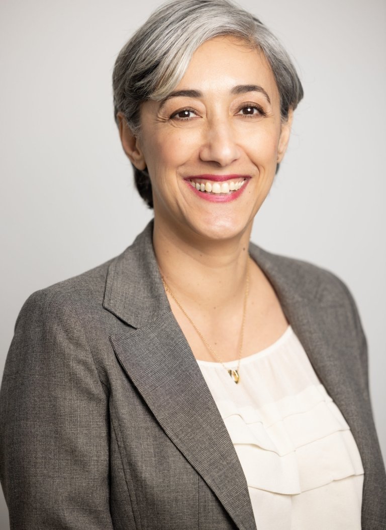 Person with short gray hair, smiling, wearing gray blazer and white shirt, in front of gray background.