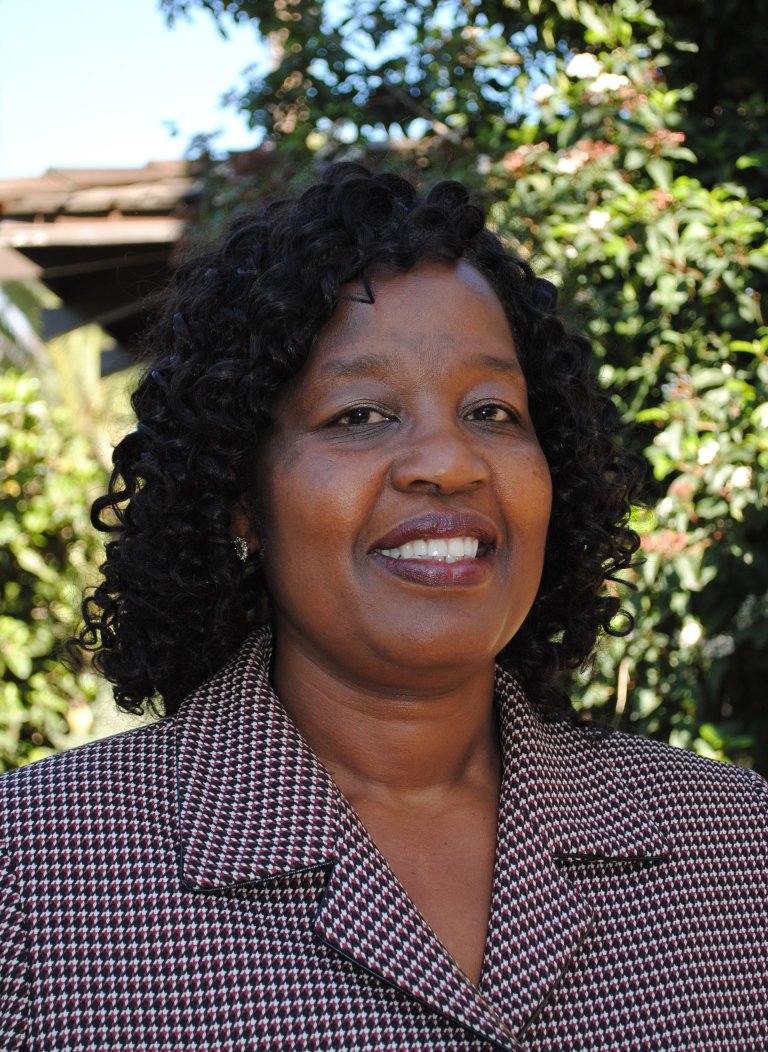 Woman, smiling, wearing collared-top, standing in front of greenery.