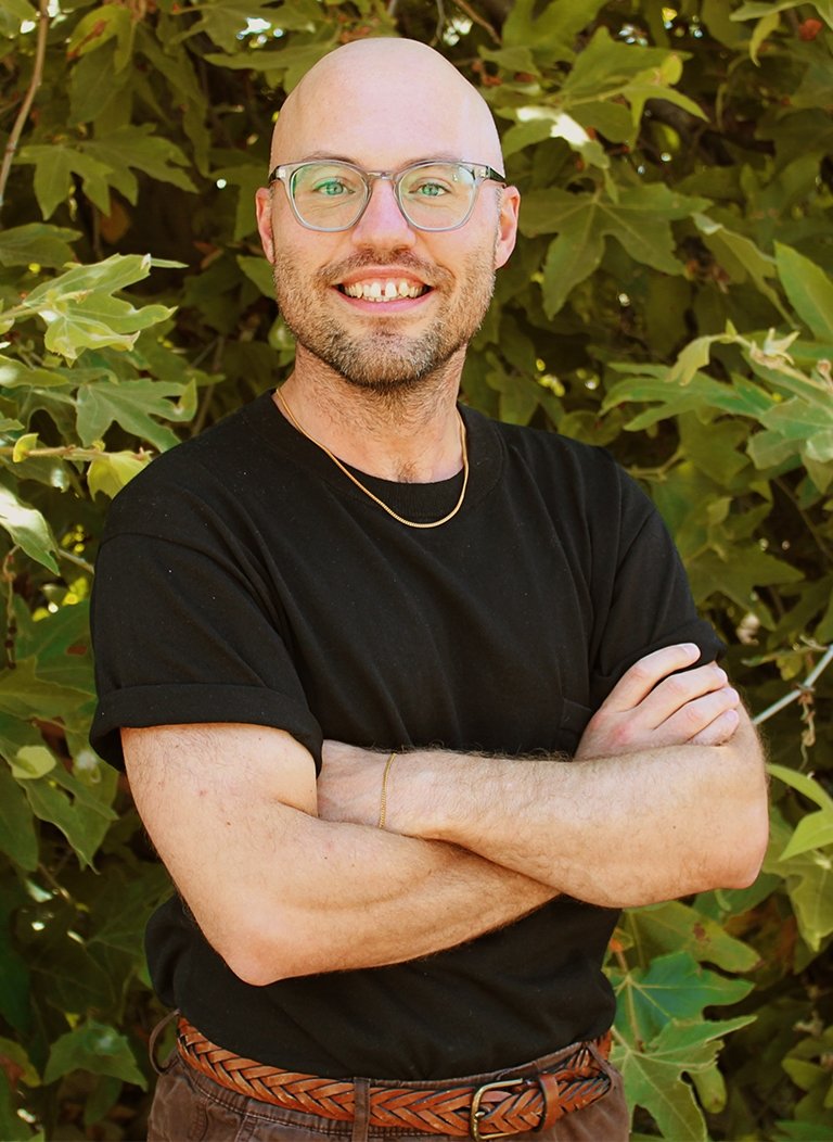 Man wearing glasses, a black shirt, and brown jeans crossing arms.