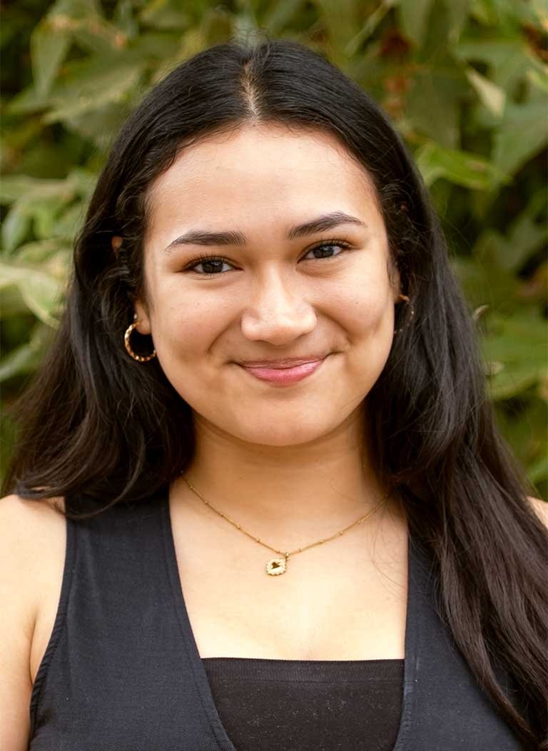 a student with long brown hair wears a black vest while standing in front of a plant on campus